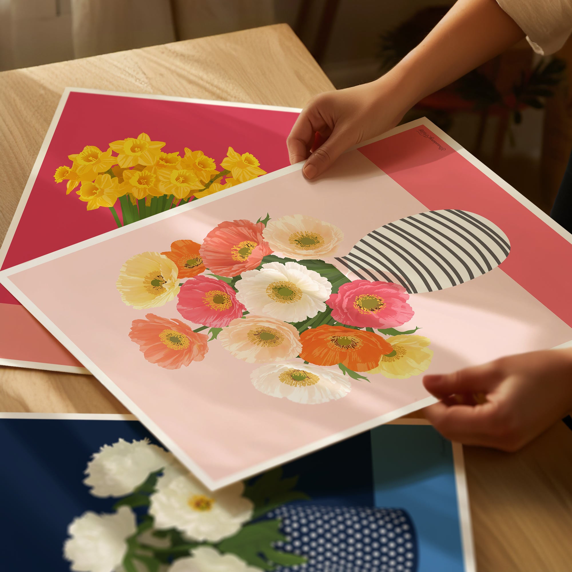 Floral art prints on a table with hands holding one of them.
