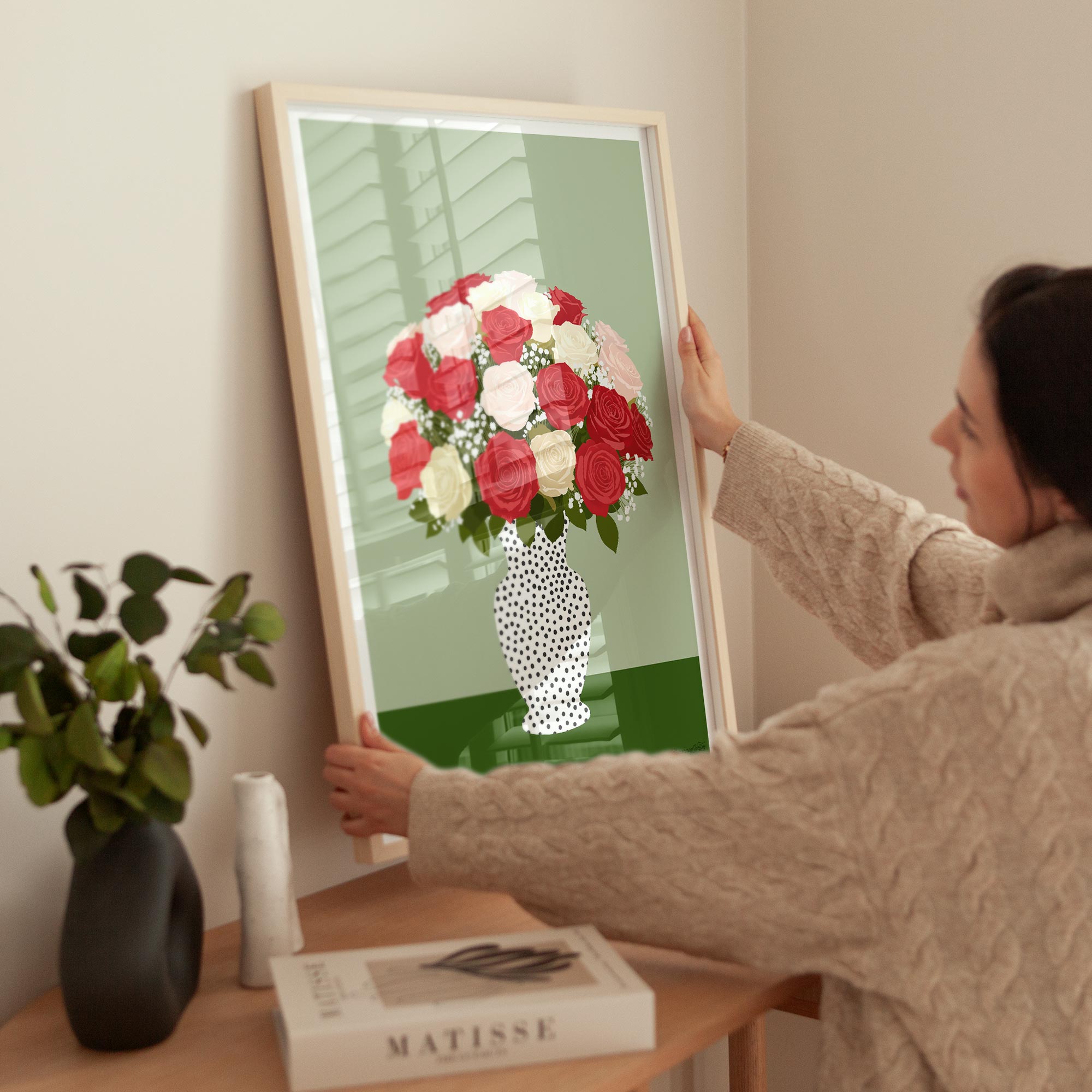 Person holding a framed artwork of a bouquet of flowers on a table with a book and plant.