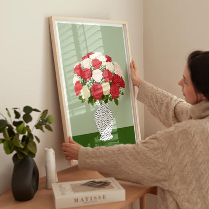 Person holding a framed artwork of a bouquet of flowers on a table with a book and plant.