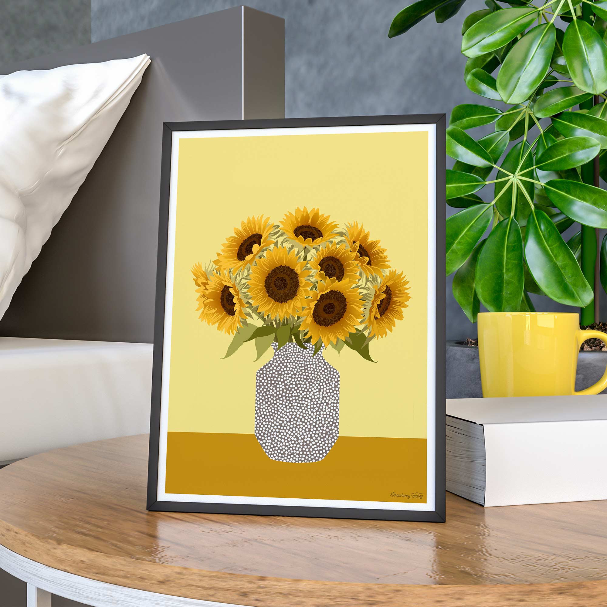 Framed artwork of sunflowers on a yellow background, placed on a wooden surface with a plant and mug in the background.