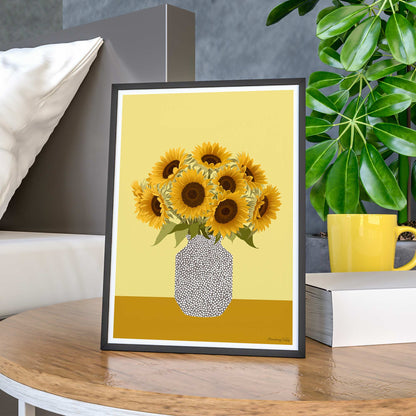 Framed artwork of sunflowers on a yellow background, placed on a wooden surface with a plant and mug in the background.